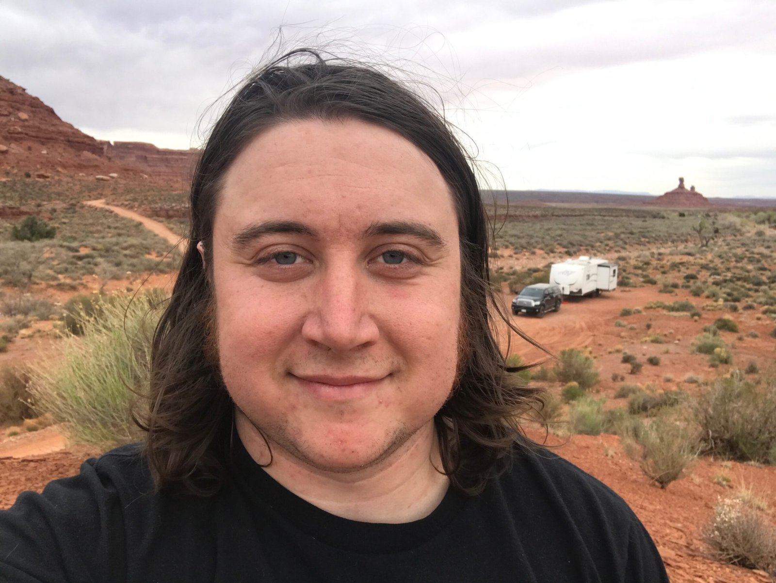 A desert landscape with me smiling in the foreground, and my RV parked in a site downhill in the distance.