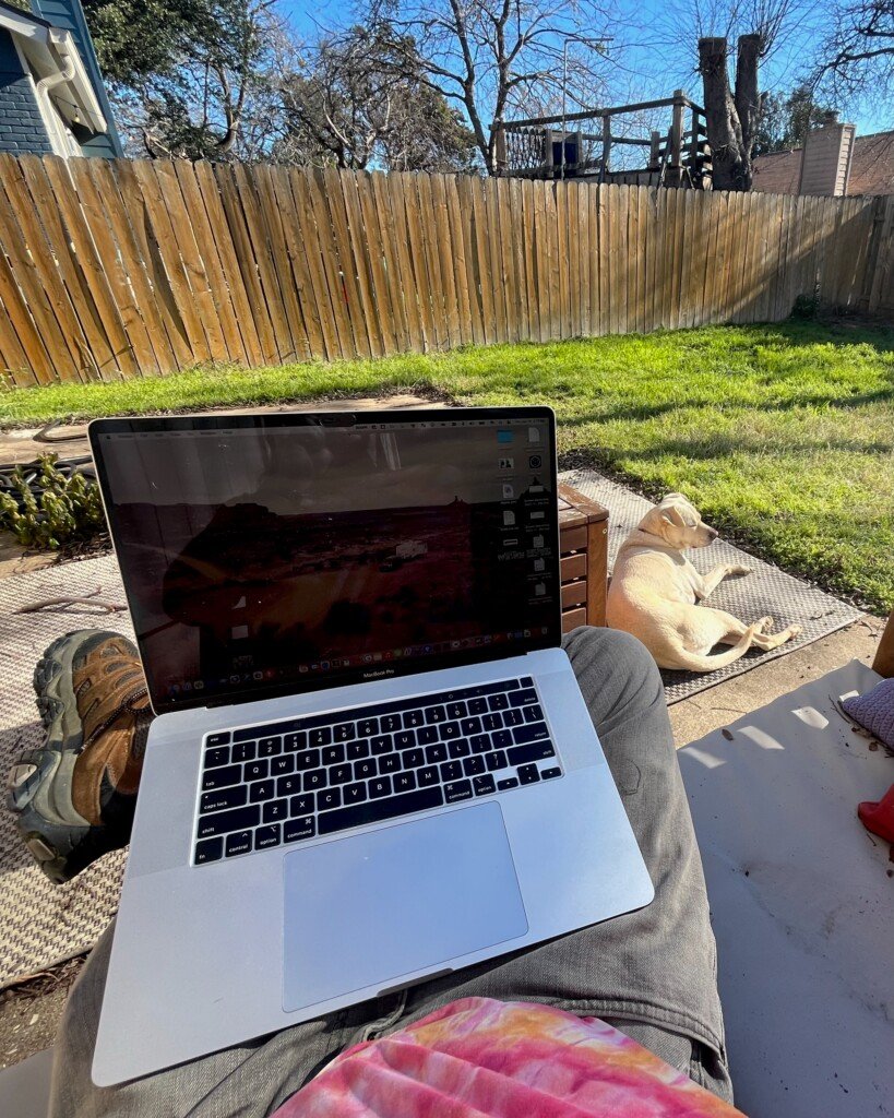 POV shot sitting on a backyard couch with a large MacBook Pro. My medium-sized dog Athena is sunbathing with eyes closed at my feet.